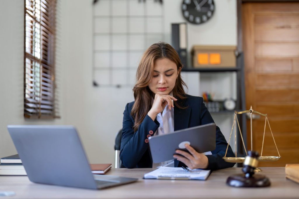 Female Attorney Working on Her Tablet and Laptop with the Scales of Justice on Her Desk